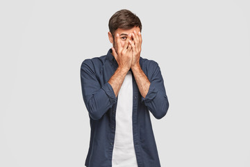 Handsome man with dark hair, bristle, hides face with hands, peeks through fingers, wears fashionable shirt, feels shy, isolated over white background. Curious guy cannot to look at present.