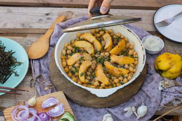 Woman hands open lid of baked chickpea with quince in cast iron skillet, frying pan. Vegan vegetarian lunch, paleo diet, healthy dinner served with salad and vegetables on table