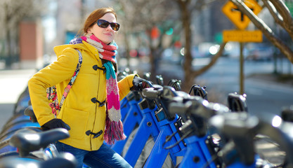Naklejka premium Happy young woman tourist ready to ride a rental bicycle in New York City at sunny spring day. Female traveler enjoying her time in downtown Manhattan.