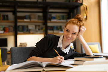 Happy redhead lady student sitting at the table with books in library writing notes.