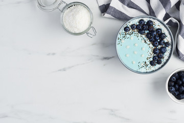 Top view of blue yogurt smoothie bowl made with blueberry, coconut flour, chia seeds and sugar pearls on white marble table with copy space.