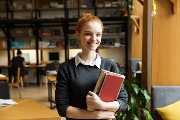 Happy redhead lady student posing indoors in library holding books.