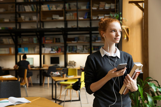 Redhead lady student posing indoors in library holding books istening music with earphones using mobile phone.