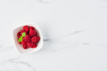Top view of fresh sweet raspberries with mint leaves in white bowl on white marble table with copy space.