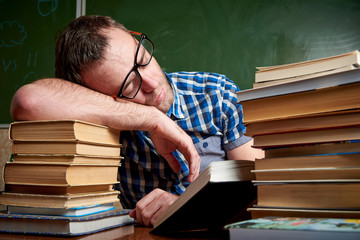 Tired and tortured disheveled student in glasses is sleeping at the table on a stack of books...