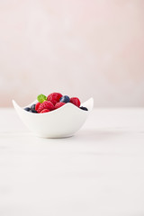 Fresh blueberries and raspberries with mint leaf in white bowl on white marble table over rose background with copy space.