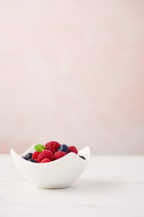 Fresh blueberries and raspberries with mint leaf in white bowl on white marble table over rose background with copy space.