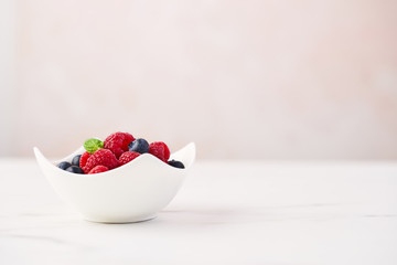 Fresh blueberries and raspberries with mint leaf in white bowl on white marble table over rose background with copy space.