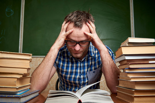 A disheveled tired unshaven young man with glasses holds his head and reads a book at the table with piles of books against the background of a blackboard.