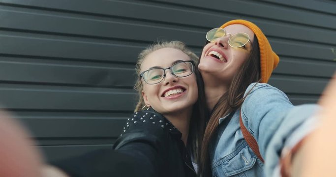 Close up of the stylish Caucasian cheerful hipster girls in taking cool selfies pictures and posing with rock and victory gestures. Outside. POV. Portrait.