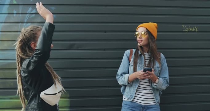 Caucasian young stylish hipster girl standing outdoor and using her smartphone when her friend with dreadlocks coming, giving her five and they both watching something on its screen.