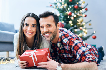 Photo of smiling couple looking at camera while relaxing in decorated room exchanging christmas present
