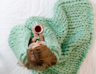 Young woman wrapped in light green merino wool blanket sitting on bed and holding cup of tea. Plaid...