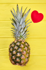 Pineapple and red heart, top view. Composition with whole ripe pineapple fruit and red felt heart on yellow wooden background.