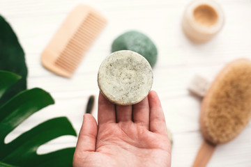 Hand holding natural solid shampoo bar on background of bamboo brush, deodorant, sponge on white wood with green monstera leaves. Zero waste. Choice plastic free eco products - obrazy, fototapety, plakaty