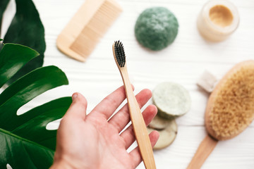Hand holding natural bamboo toothbrush on background of shampoo bar, brush, toothpaste on white wood with green monstera leaves. Zero waste. Choose plastic free eco products