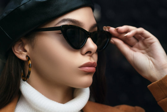 Outdoor close up fashion portrait of young beautiful woman wearing cat eye sunglasses, leopard print hoop earrings, leather beret. Model looking aside