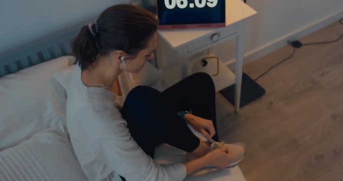 Attractive sporty young Caucasian woman getting ready for exercising early in the morning, tying her sports shoe and choosing music on her phone. 4K UHD