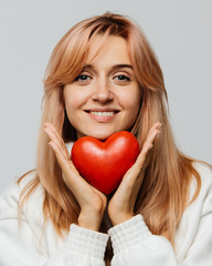 Portrait of joyful attractive woman with strawberry blonde hair hold red heart (Valentine day symbol),looking at camera, closeup, isolated on light background.Love, happiness, relationships concept