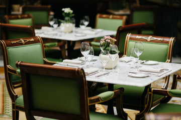 Table served with luxury glasses, cutlery, flowers, green chairs around in cozy restaurant. Nobody in shot. Decorated table for festive event