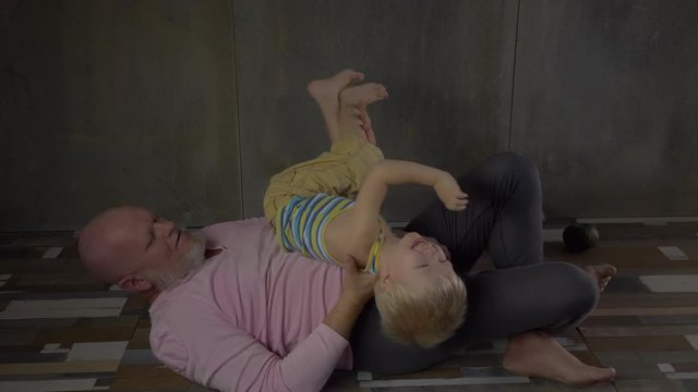 elderly man in pink sweater with gray beard and shaved head doing physical exercises on floor. Senior instead of dumbbells raises little blond boy. concept of friendly family