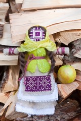 motanka, Ukrainian ethnic traditional toy, handmade textile rag doll, folk craft souvenir, shawl, embroidery, canvas, wool, stands on bark with apple before firewood