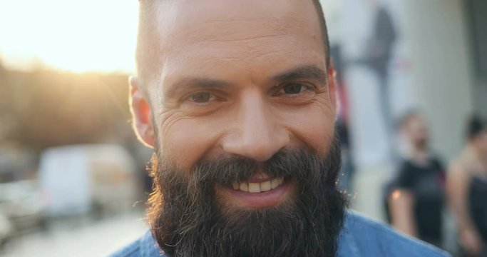 Portrait of the Caucasian attractive man with a beard looking straight to the camera with a shrewd eyes and then smiling sincerely. Close up of the face. Outdoor.