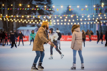 Fototapeta na wymiar Young couple in love Caucasian man with blond hair with long hair and beard and beautiful woman have fun, active date skating on ice scene in town square in winter on Christmas Eve