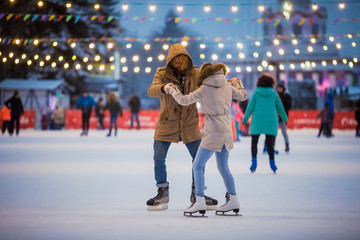 Young couple in love Caucasian man with blond hair with long hair and beard and beautiful woman have fun, active date skating on ice scene in town square in winter on Christmas Eve - obrazy, fototapety, plakaty