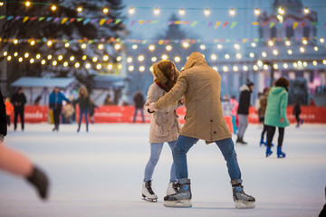 Young couple in love Caucasian man with blond hair with long hair and beard and beautiful woman have fun, active date skating on ice scene in town square in winter on Christmas Eve