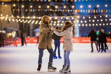 Young couple in love Caucasian man with blond hair with long hair and beard and beautiful woman have fun, active date skating on ice scene in town square in winter on Christmas Eve