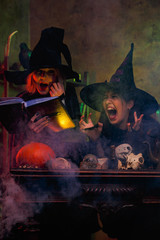 Image of laughing two witches in black hats reading book of incantations on cauldron with steam