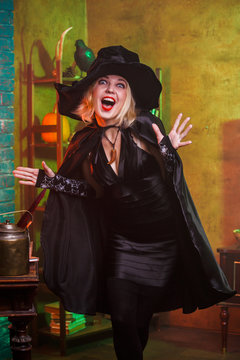 Photo of happy witch in black hat, dress on background of rack with pumpkin and crow