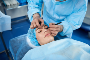 Calm lady lying with her eyes closed before the plastic surgery and the doctor drawing lines on her face