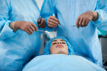 Close up of young lady lying with her eyes closed and professional surgeons holding medical instruments near her face - obrazy, fototapety, plakaty