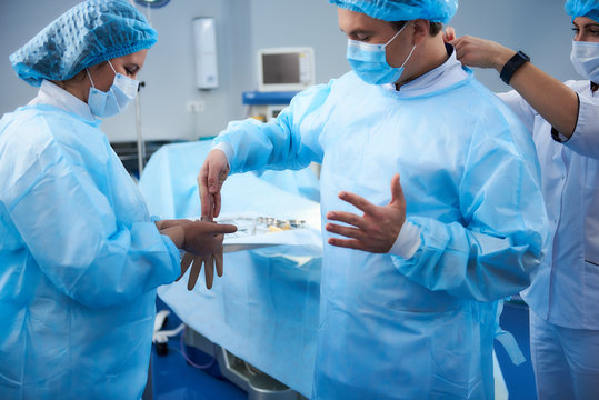 Helpful Young Nurse Holding Rubber Gloves And Serious Surgeon Putting Them On Before The Surgery