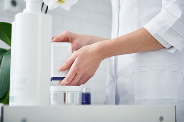 Close up of female hands with cosmetic and personal care products on table. Lady is wearing white lab coat