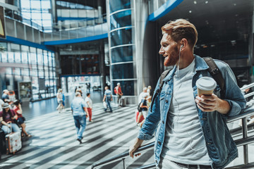 Waist up of a pleasant bearded casual man drinking coffee while standing at the railway station
