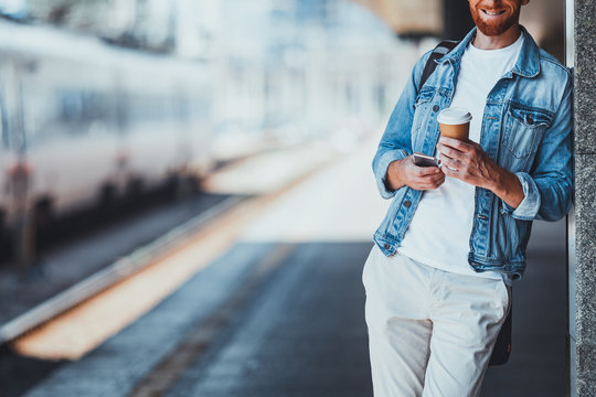 Cheeful smiling young man standing atthe railway station while enjoying tasty coffee