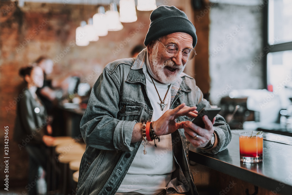 Wall mural waist up portrait of cheerful hipster pensioner sitting in bar and using smartphone