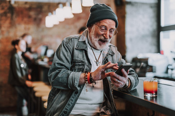 Waist up portrait of cheerful hipster pensioner sitting in bar and using smartphone