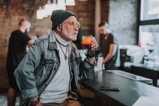 Waist up portrait of thoughtful hipster pensioner sitting in bar and drinking cocktail while looking in window