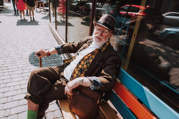 Full length portrait of good looking smiling senior male resting on colorful bench and holding stick