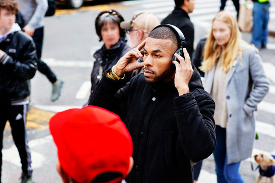 Portrait of a young man listening and holding headphones while crossing the street amongst crowd