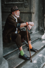 Good looking old man with white beard and cheerful smile sitting on stairs of city building