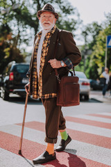 Concept of positive mood in old age lifestyle. Full lenght portrait of hipster smiling senior male going around the city with stick and briefcase