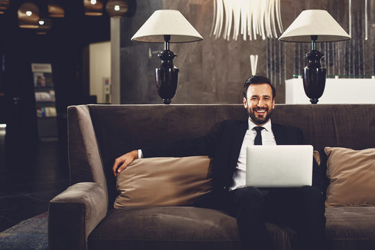 Positive bearded man smiling and sitting comfortably on the sofa while holding modern laptop