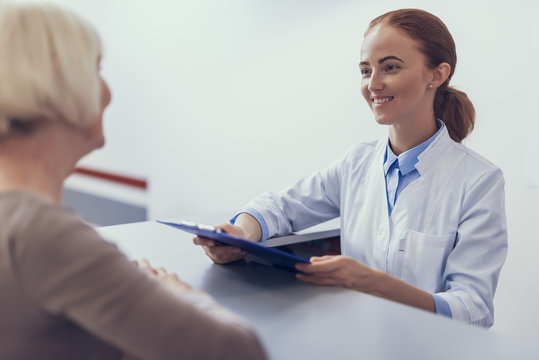 Focus on smiling female practitioner standing at reception desk. She is holding clipboard and looking at woman standing in front. Physician is meeting visitor in lobby