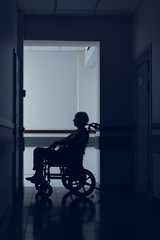 Fototapeta na wymiar Woman is using wheelchair in clinic corridor. She is sitting in solitude in dark passageway and waiting for doctor