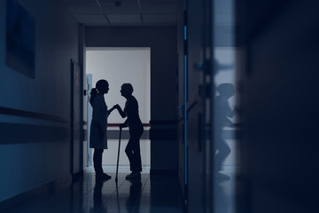 Woman and physician are standing in hospital corridor. Patient is walking with stick while female practitioner is holding her hand. Copy space in right side